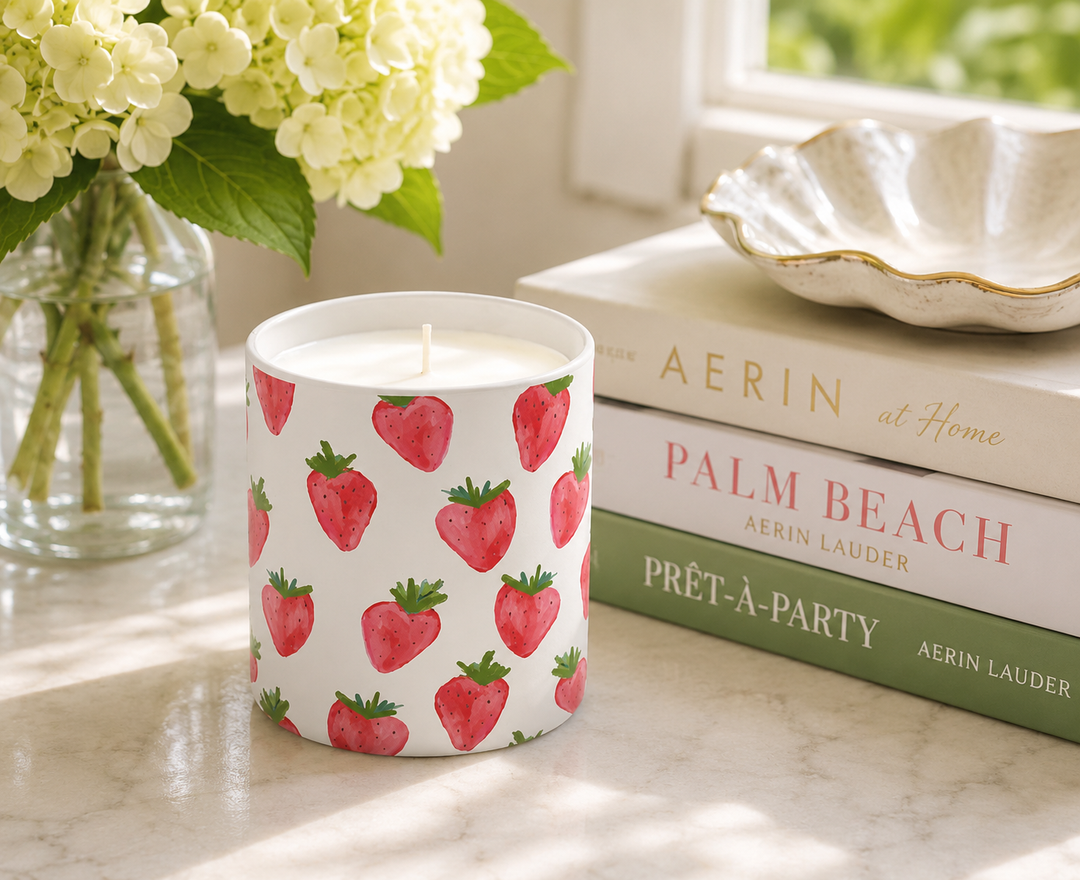 A white candle jar printed with a repeating red strawberry pattern, styled on a marble surface next to a vase of white flowers and a stack of books.