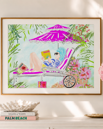 A framed art print featuring a colorful illustration of a woman lounging on a pink chaise lounge under a purple umbrella, styled on a white console table with books and a vase of pink flowers.