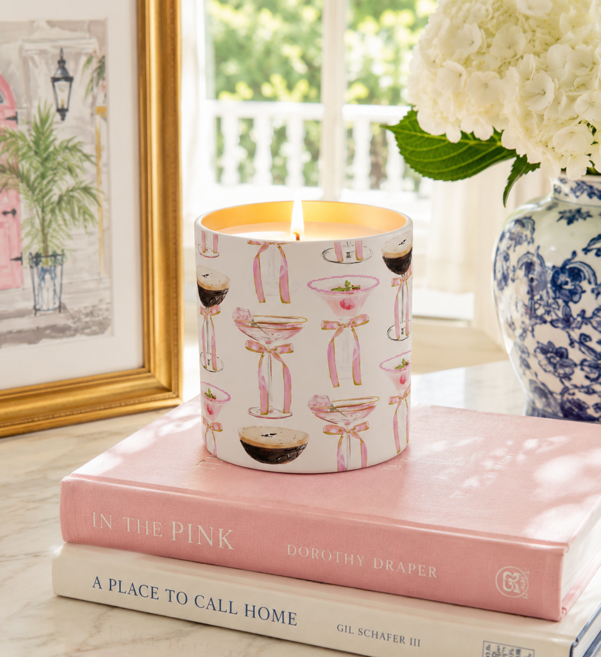 A lit white candle with a pink and black cocktail-themed print sits on top of two pink books on a marble surface, with a framed print, a blue-and-white vase of flowers, and a window in the background.