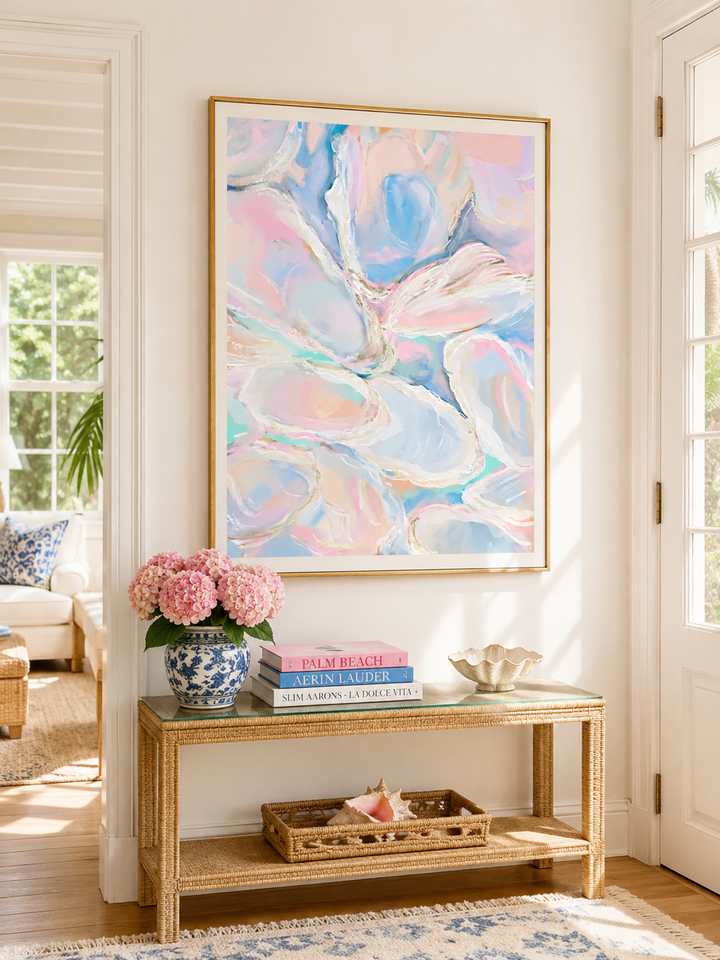 A large framed abstract painting with soft pastel blue, pink, and white brushstrokes hangs on a cream wall above a light wood console table styled with pink flowers, books, and a shell bowl.