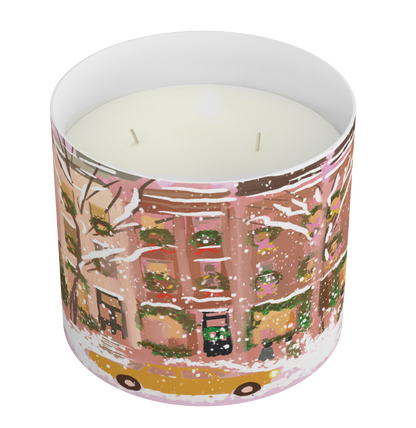A two-wick scented candle in a cylindrical glass container with a winter cityscape illustration. The artwork features pink and brown building facades, snow-covered trees, green wreaths, and a yellow taxi at the bottom, set against a plain white background.