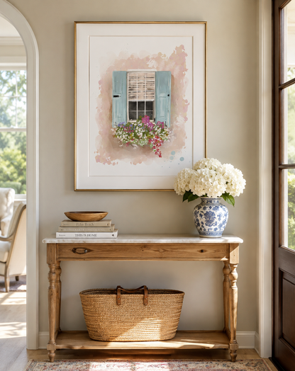 A framed art print depicting a watercolor window with blue shutters and pink flowers, displayed on a beige wall above a wooden console table with a vase of white flowers and a woven basket.