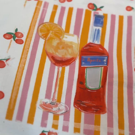 White t-shirt with a hand-painted Aperol spritz cocktail illustration, orange slice garnish, striped background, and scattered citrus details, designed by Rebecca Illustrated.