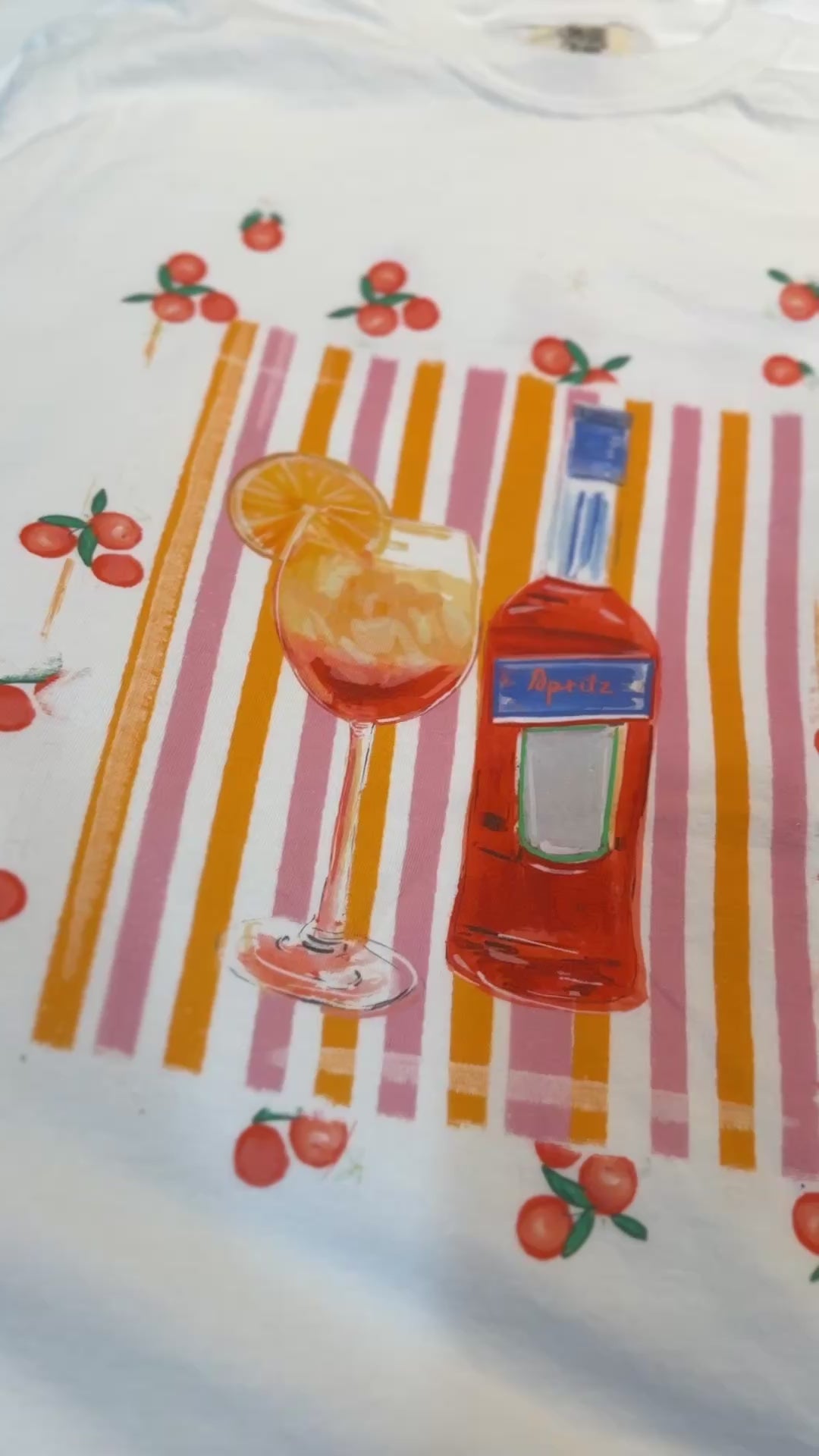 White t-shirt with a hand-painted Aperol spritz cocktail illustration, orange slice garnish, striped background, and scattered citrus details, designed by Rebecca Illustrated.