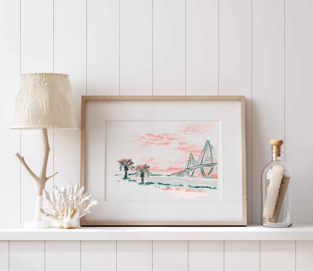 Art print of the Ravenel Bridge and palmetto trees under a pink sunset sky, painted in soft coastal colors and framed on a white wall.